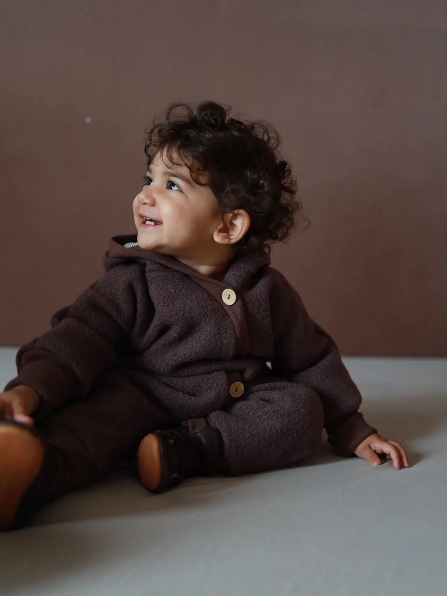 A toddler wears a Mauve wool-fleece romper with a hood and wooden buttons, made of 100% virgin wool with a cotton lining, sitting on the floor with a smile, demonstrating freedom of movement and warmth.