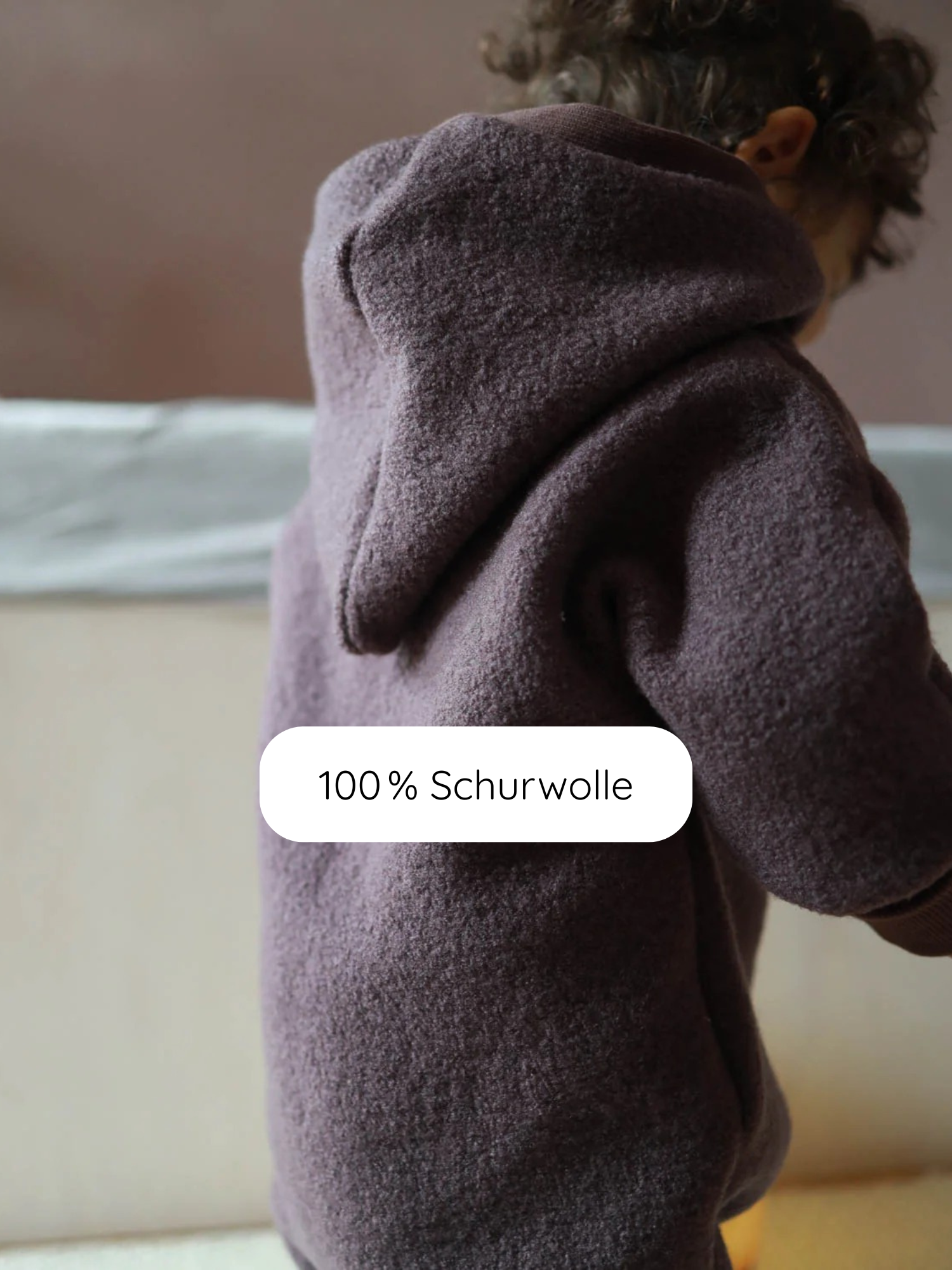Kleinkind von hinten im Kuschelanzug Wollwalk - Malve aus 100 % Schurwolle mit Kapuze, zeigt den flauschigen Stoff. Überlagert ist der Schriftzug „100 % Schurwolle“.
