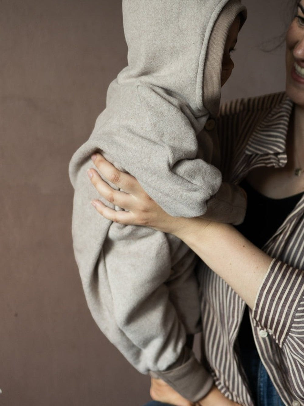 petit cochon Baby Overall aus beigem Bio-Baumwollfleece mit Wichtelkapuze, gehalten von einer Mutter