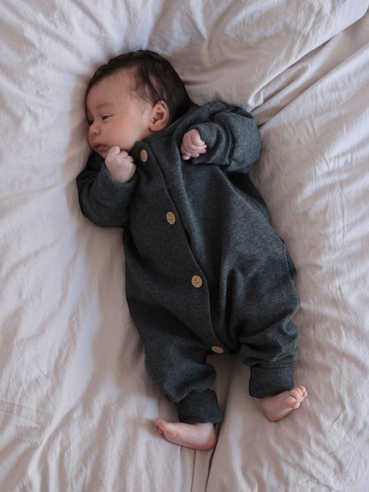 Baby in einem petit cochon Mini Overall aus dunkelgrauem, meliertem Feinstrick-Jersey mit Holzknöpfen auf dem Bett.