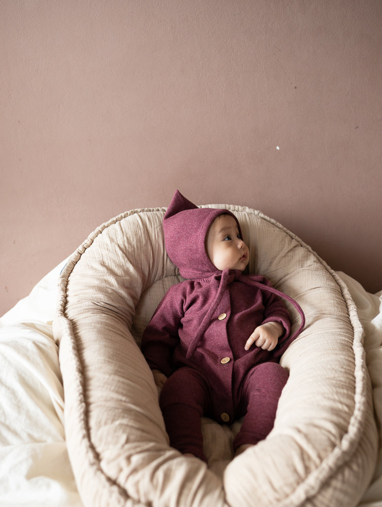 Baby in einem weinroten petit cochon Mini Overall aus meliertem Feinstrick mit Kapuze in einem beigen Babynest