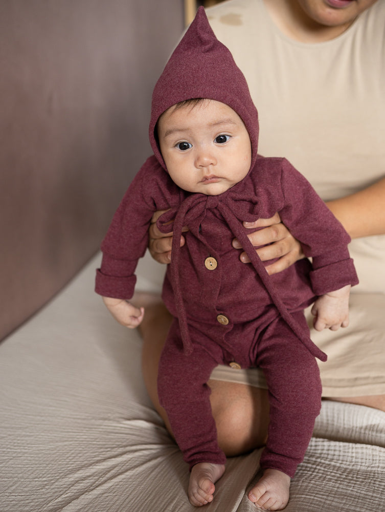 Baby trägt einen weinroten petit cochon Mini Overall aus meliertem Feinstrick mit Kapuze und Holzknöpfen