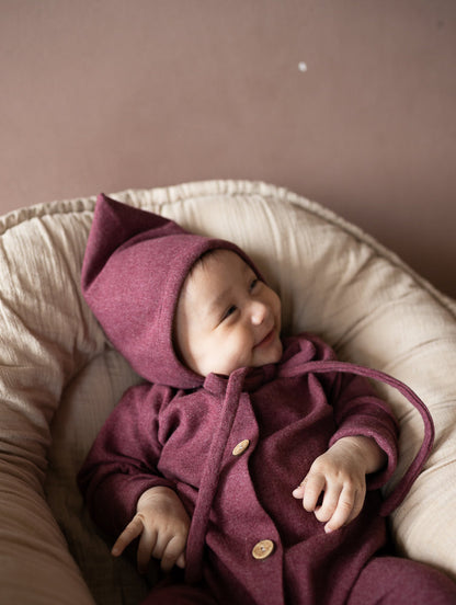 Baby in einem weinroten petit cochon Mini Overall aus meliertem Feinstrick mit passender Zipfelmütze im Babynest