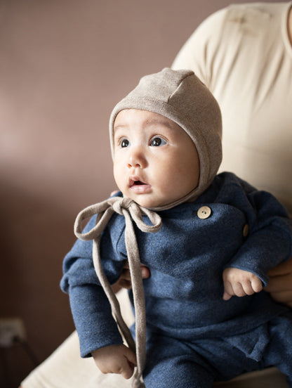 Baby trägt blaues petit cochon Wickeloberteil aus Feinstrick mit Kokosknöpfen und eine passende beige Strickhaube
