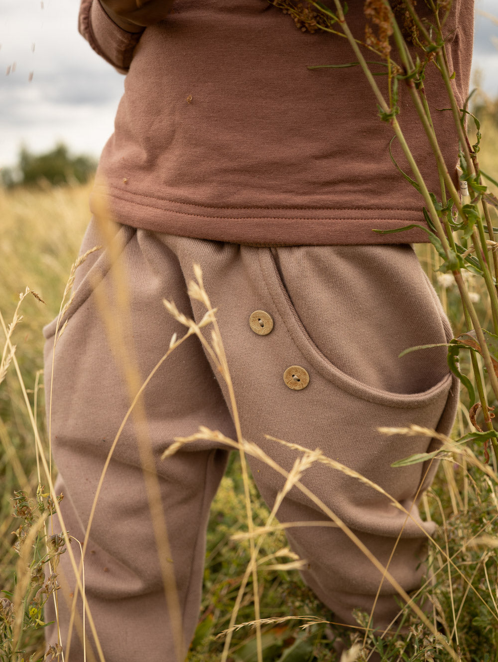 petit cochon Knickerbocker Hose aus schokobraunem Bio-Sweat mit Kokosknöpfen, getragen auf einer Wiese
