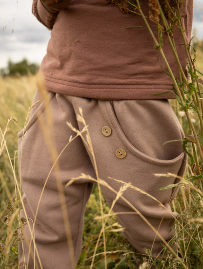 petit cochon Knickerbocker Hose aus schokobraunem Bio-Sweat mit Kokosknöpfen, getragen auf einer Wiese