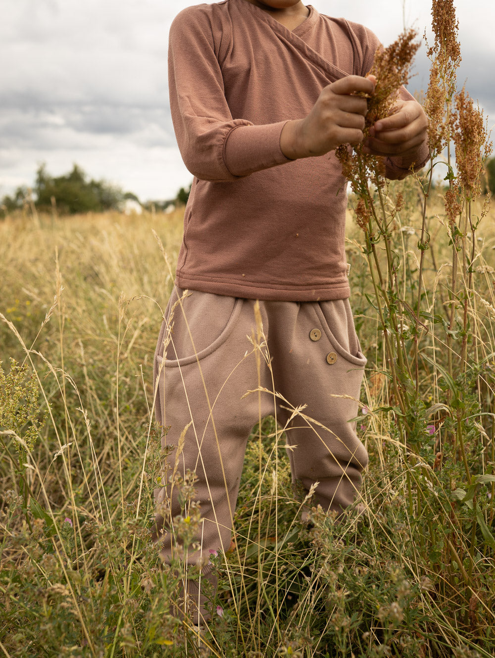 petit cochon Knickerbocker Hose aus schokobraunem Bio-Sweat mit Kokosknöpfen, getragen von einem Kind auf einer Wiese.