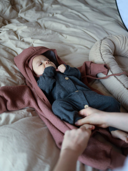 petit cochon Mini Overall aus meliertem Feinstrick in Dunkelgrau mit Kapuze, getragen von einem Baby im Bett