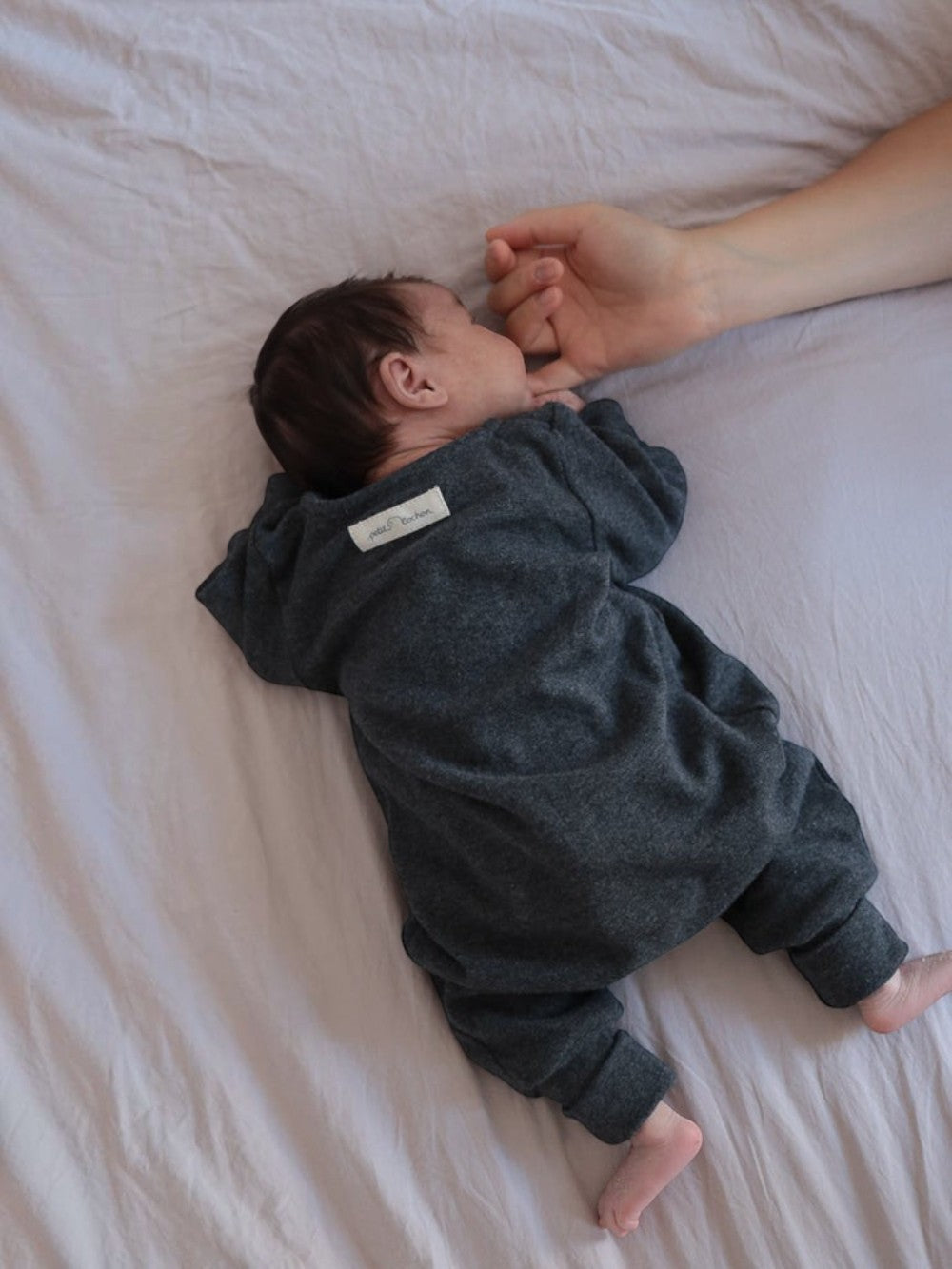 petit cochon Mini Overall aus dunkelgrauem meliertem Feinstrick-Jersey, getragen von einem Baby im Bett