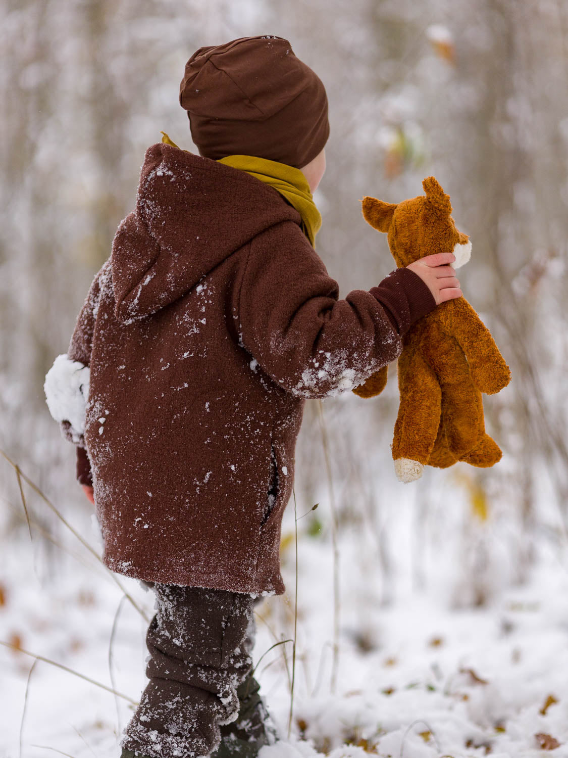 Kind in brauner petit cochon Wollwalk-Wichteljacke mit Kapuze und Bio-Jersey-Futter im verschneiten Wald