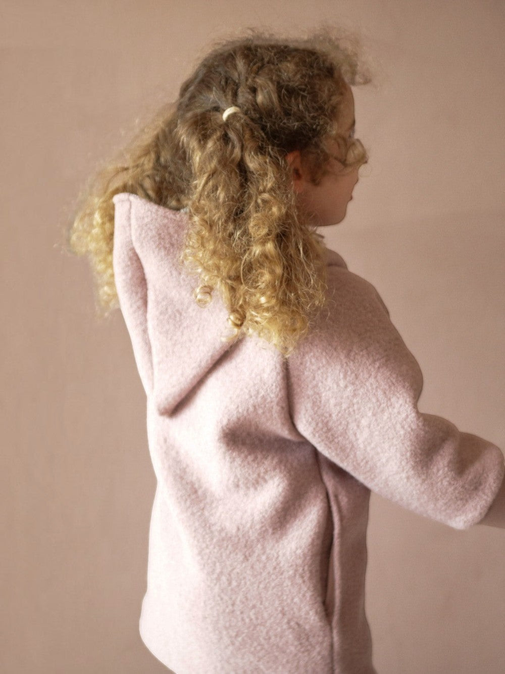 petit cochon Wichteljacke aus rosa Wollwalk mit Kapuze, getragen von einem Kind mit lockigem Haar
