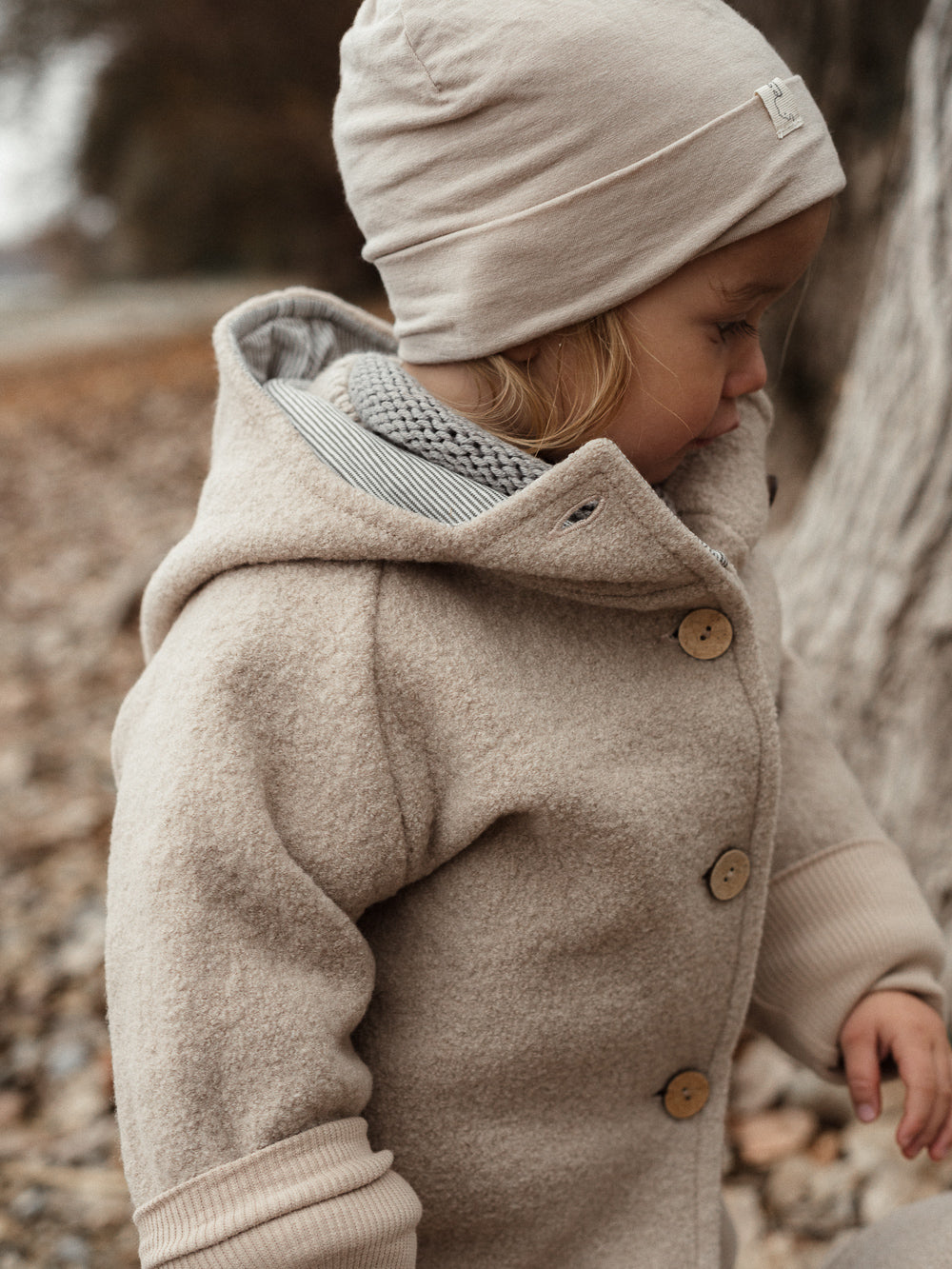 petit cochon Wichteljacke aus beigem Wollwalk mit Kapuze, Bio-Jersey-Futter und Holzknöpfen an einem Kind im Freien
