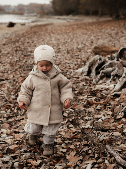 petit cochon Wichteljacke aus beigem Wollwalk mit Bio-Jersey-Futter, getragen von einem Kind im Herbstlaub.