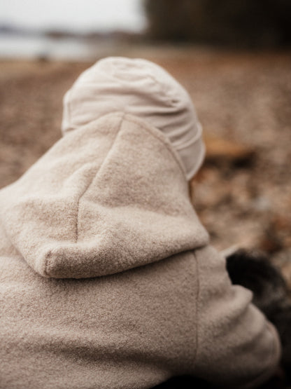 petit cochon Wichteljacke aus beigem Wollwalk mit Kapuze, getragen von einem Kind in natürlicher Umgebung