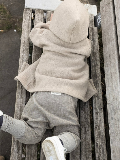 petit cochon Wichteljacke aus beigem Wollwalk mit Kapuze, getragen von einem Kind auf einer Holzbank im Freien