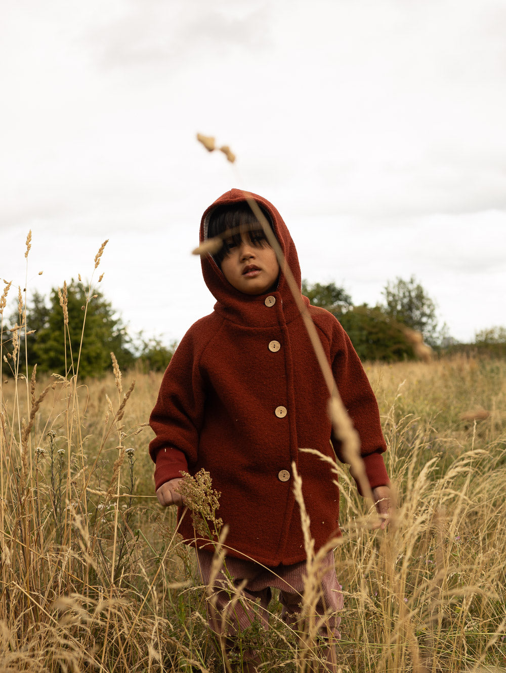 Kind trägt eine rostrote petit cochon Wichteljacke aus Wollwalk mit Kapuze und Knöpfen auf einem herbstlichen Feld