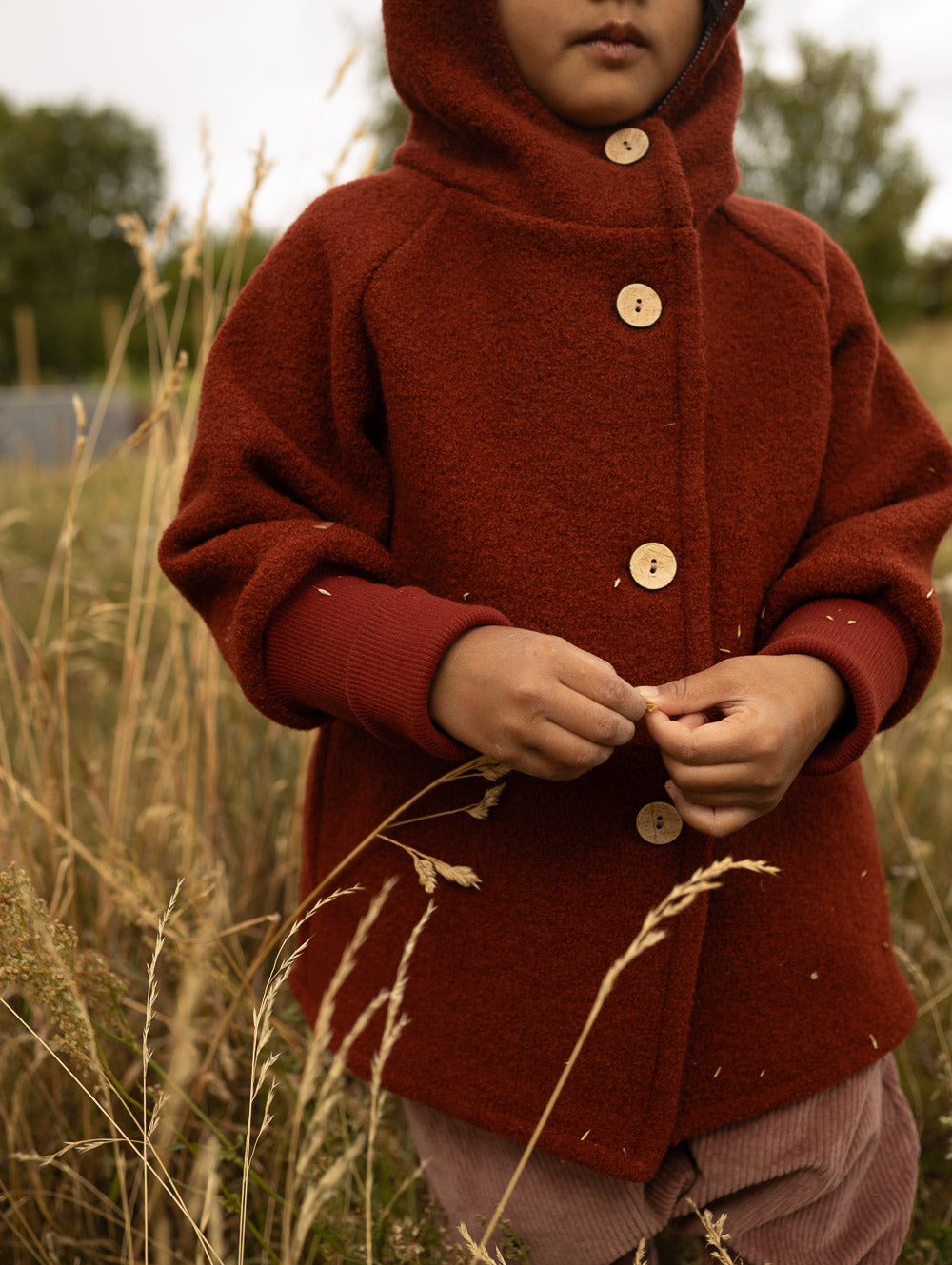 petit cochon Wichteljacke aus rostrotem Wollwalk mit Kapuze, Holzknöpfen und Bio-Jersey-Bündchen im Freien