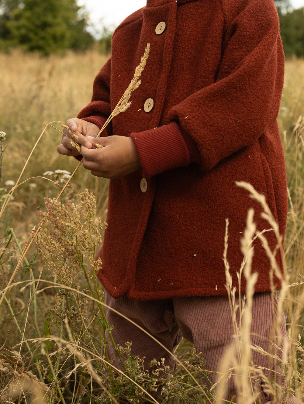 Kind in einer rostroten petit cochon Wichteljacke aus Wollwalk mit Holzknöpfen und Bündchen auf einem Feld