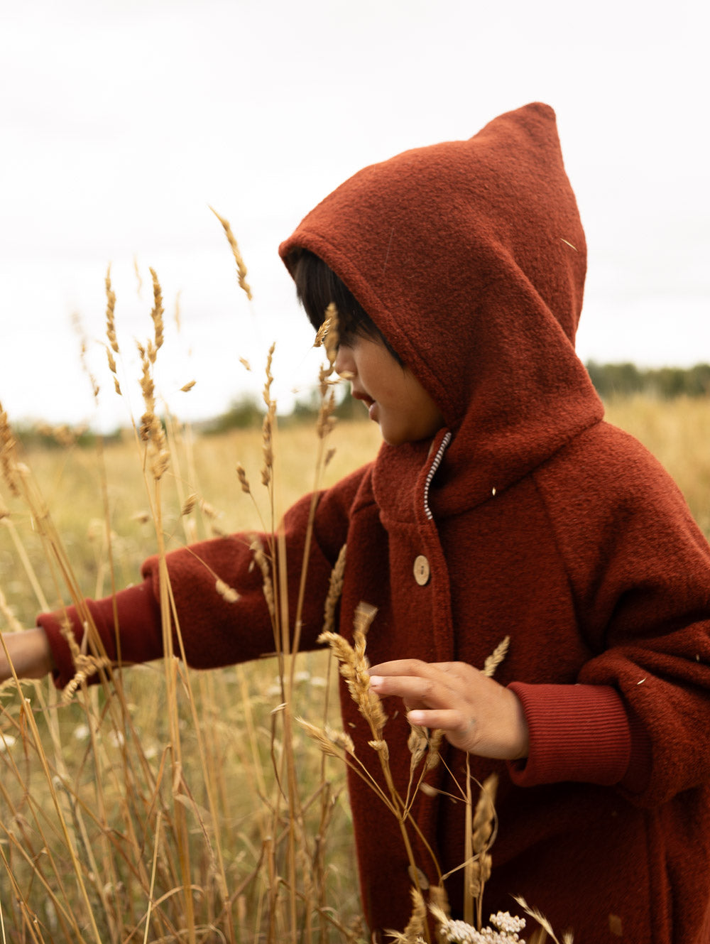 Kind trägt eine rostrote petit cochon Wichteljacke aus Wollwalk mit Kapuze beim Spielen in einem herbstlichen Feld