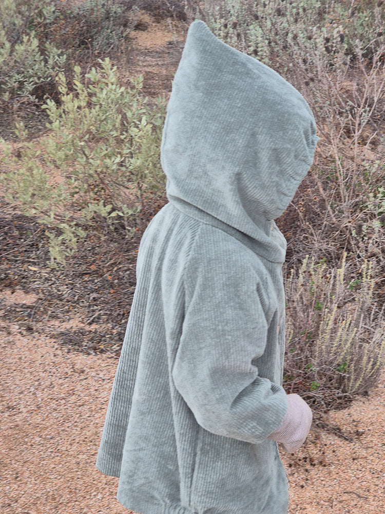 Ein Kind trägt die Wichteljacke Breitcord Stretch-Aqua, eine locker sitzende Jacke mit Wichtelkapuze aus sagegrünem Cord, auf einem sandigen Pfad.