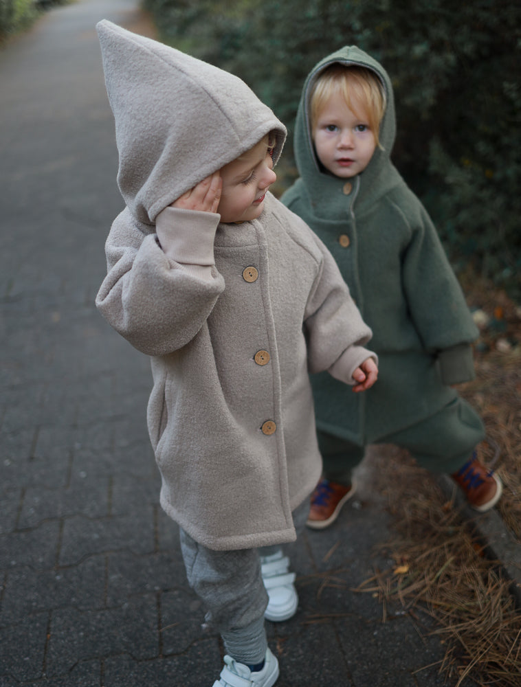 Zwei Kinder in Wichteljacken Wollwalk - Sand und Grün, warm eingepackt mit Kapuzen, stehen auf einem Weg im Freien. Die Jacken sind aus Wollwalk mit Bio-Jersey-Futter, ideal für kühle Jahreszeiten.