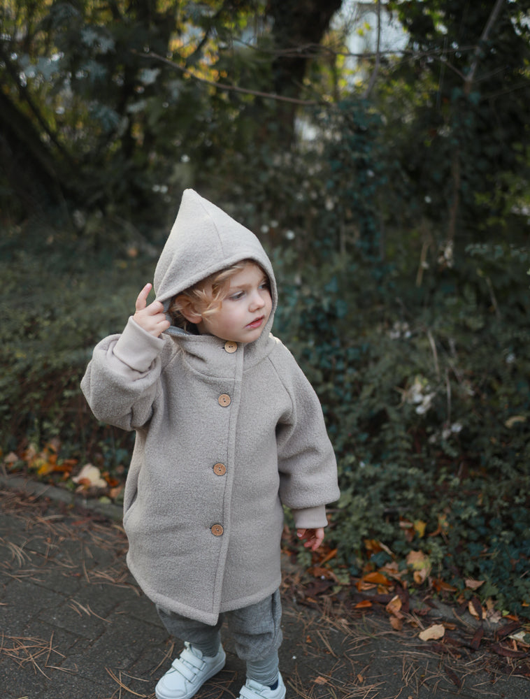Ein Kind trägt die Wichteljacke Wollwalk - Sand mit Kapuze und Holzknöpfen. Die Jacke bietet Bewegungsfreiheit und Komfort, ideal für Herbst und Winter.