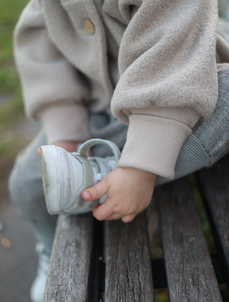 Ein Kind auf einer Parkbank zieht einen Schuh an und trägt die Wichteljacke Wollwalk - Sand. Die Jacke bietet Bewegungsfreiheit und ist ideal für kühlere Jahreszeiten.