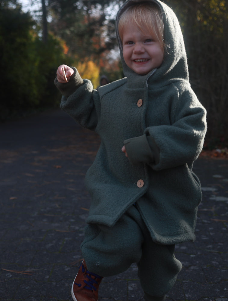 Kleinkind in Knickerbocker Wollwalk - Pistazie, lächelnd in grüner Kapuzenjacke und Hose. Hält ein Blatt, im Gehen auf einem Weg. Zeigt die robusten, wachsenden Krempelbündchen der Hose.