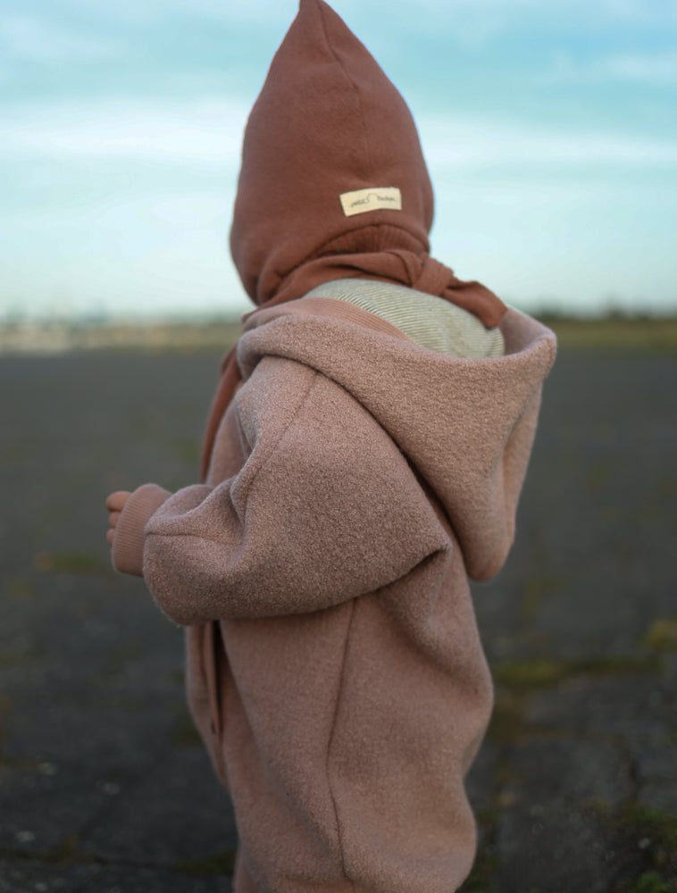 Ein kleines Kind trägt den Kuschelanzug Wollwalk - Puder auf einem leeren Flugfeld. Der Anzug ist weich, mit Kapuze und passender Mütze, ideal für kalte Tage.