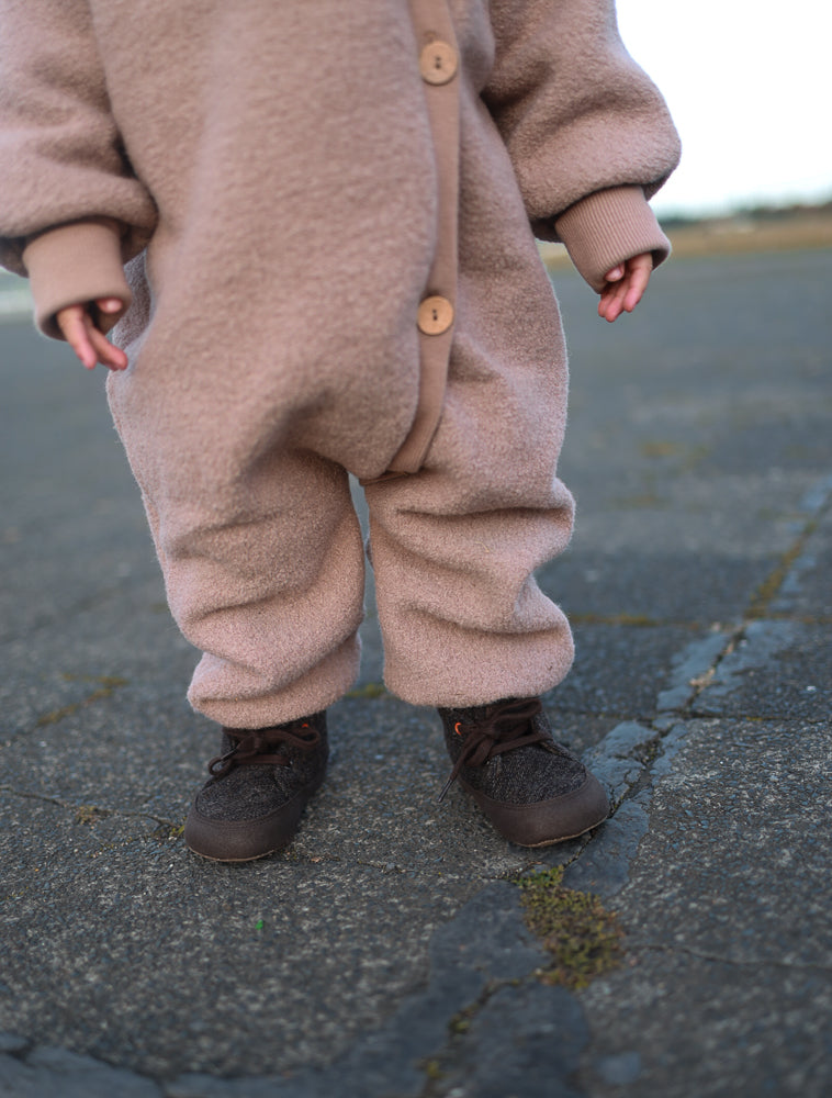 Kleinkind in Kuschelanzug Wollwalk - Puder, einem pink-beige Fleece-Einteiler mit Knöpfen und dunklen Schuhen, stehend auf Asphalt. Ideal für kühle Jahreszeiten, gefertigt aus 100% Schurwolle mit Baumwollfutter.