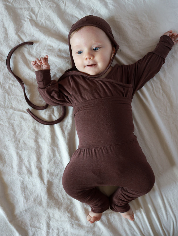 Ein Baby liegt in einem weichen, schokoladenbraunen Babyknicker Bambusviskose Zartbitter mit Kapuze und ausgestreckten Armen auf einem hellen Laken, wirkt entspannt und aufmerksam.