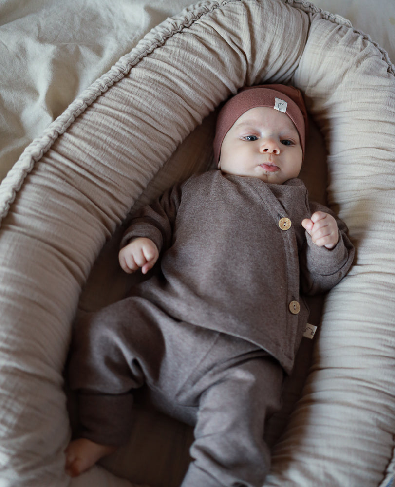 Ein Säugling liegt in einem weichen, gepolsterten Baby-Lounger und trägt das Babyschlüttli Feinstrick - Praline Meliert, bestehend aus einem warmen, langärmligen Oberteil, Hose und passender Mütze.
