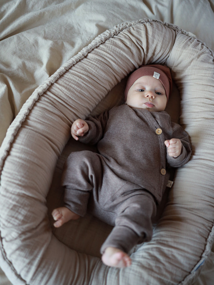 Babyschlüttli Feinstrick - Praline Meliert: Ein Baby liegt in einem weichen Nest, trägt einen braunen Einteiler mit Seitenschließe aus meliertem Feinstrick-Jersey, passend für sanften Komfort und einfache Handhabung.