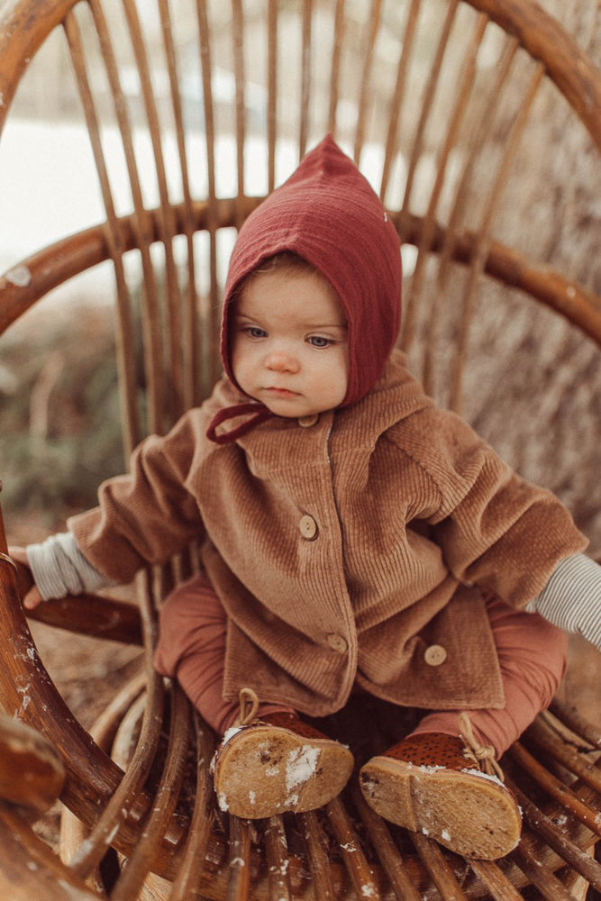 Kleinkind sitzt in einem niedrigen Korbsessel, trägt die Wichteljacke Breitcord Stretch - Weizen mit Holzknöpfen und Ringel-Armbündchen, kombiniert mit einer rostfarbenen Hose und braunen Lederschuhen.