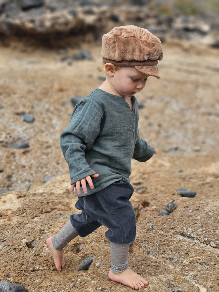 Ein Kleinkind trägt die Michelmütze Breitcord Stretch - Weizen, eine weiche, verstellbare Cordkappe mit kleinem Schirm, während es barfuß über einen steinigen Strand geht.