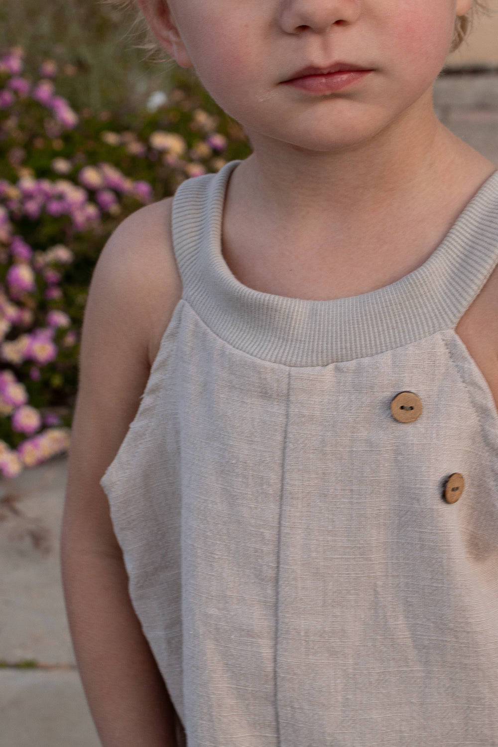 Nahaufnahme eines Kindes in einem ärmellosen, hellbeigen Oberteil mit Holzknöpfen. Im Hintergrund sind Blumen erkennbar. Dies betont die vielseitige Schlupflatzhose Leinen - Sand.