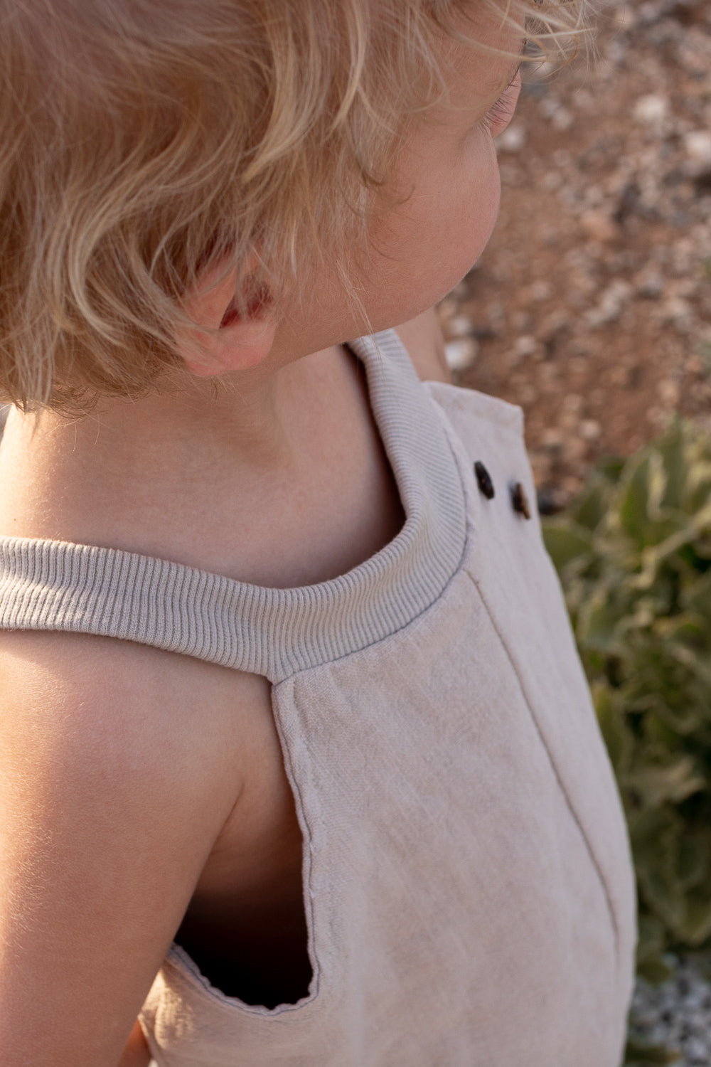 Kleinkind in einer Schlupflatzhose Leinen - Sand. Die ärmellose Hose hat einen gerippten Ausschnitt und Knopfdetails, perfekt für Spiel und Bewegung.