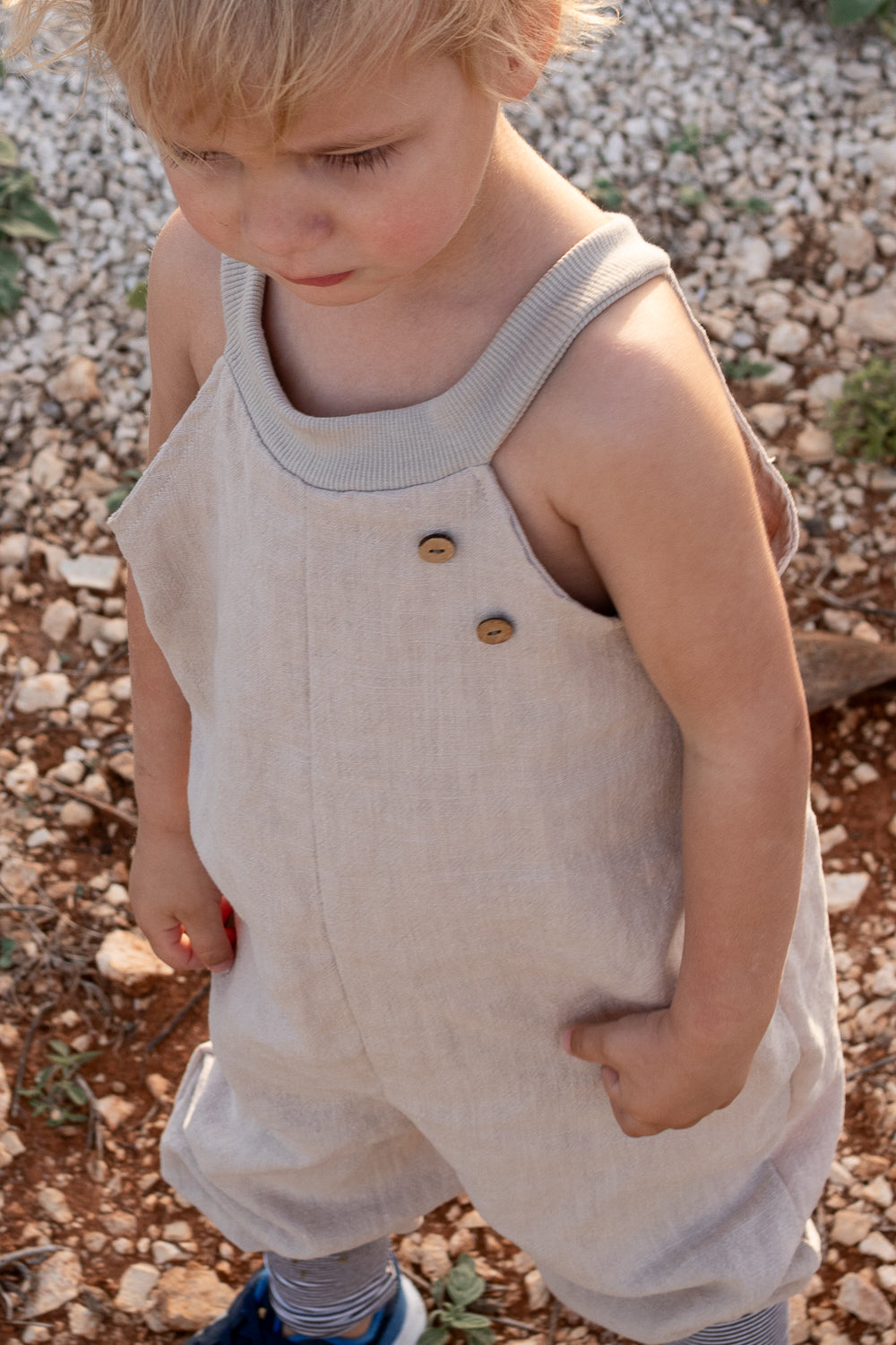 Kleinkind trägt Schlupflatzhose Leinen - Sand mit Holzknöpfen, über gestreiften Leggings und blauen Schuhen, steht auf steinigem Boden, hält etwas in der Hand, schaut nach unten.