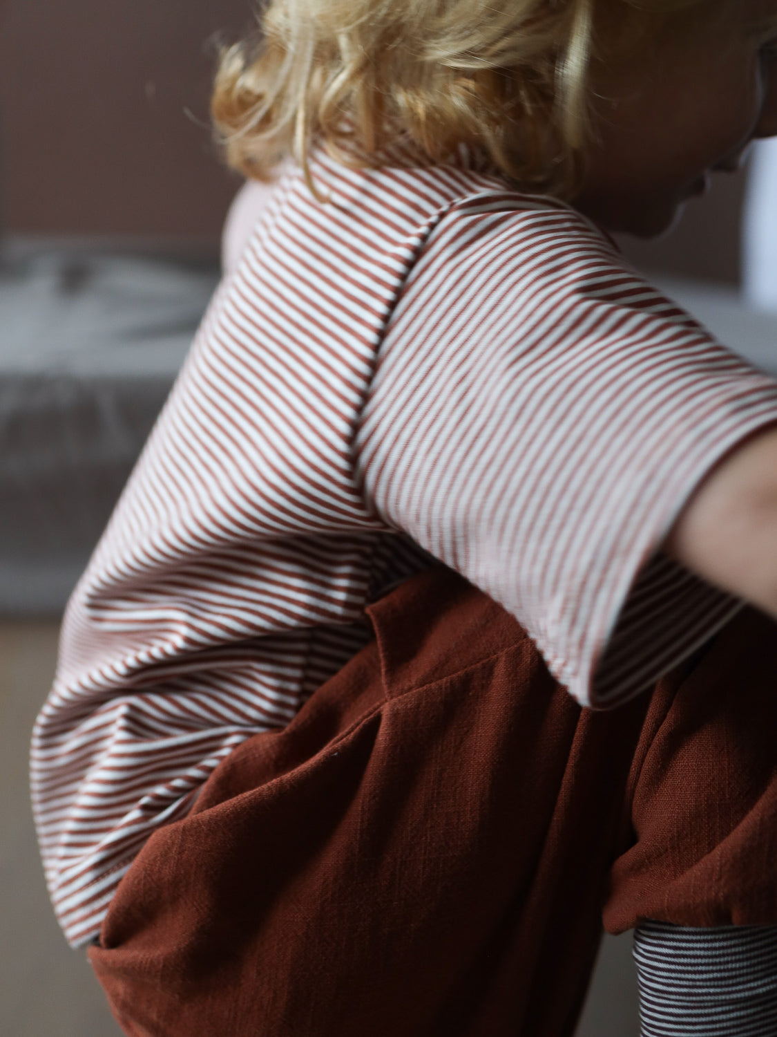 Ein Kind trägt ein kurzes Wickelshirt aus Baumwolle - Ringel Ziegel, mit schmalen roten und weißen Streifen, kombiniert mit rostfarbenen Hosen. Es ist sichtbar bequem und handgemacht.