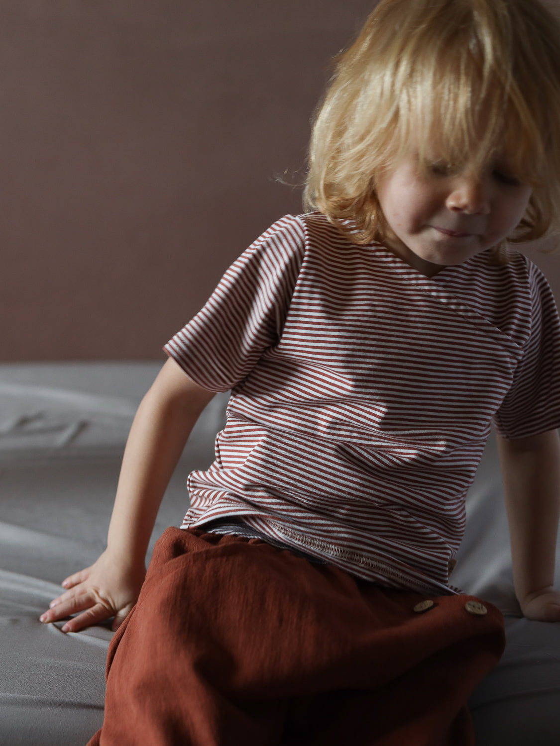 Ein Kleinkind trägt das Kurzes Wickelshirt Baumwolle - Ringel Ziegel, ein rot-weiß gestreiftes, kurzärmeliges Oberteil. Es sitzt auf einem Bett und trägt rostfarbene Hosen.