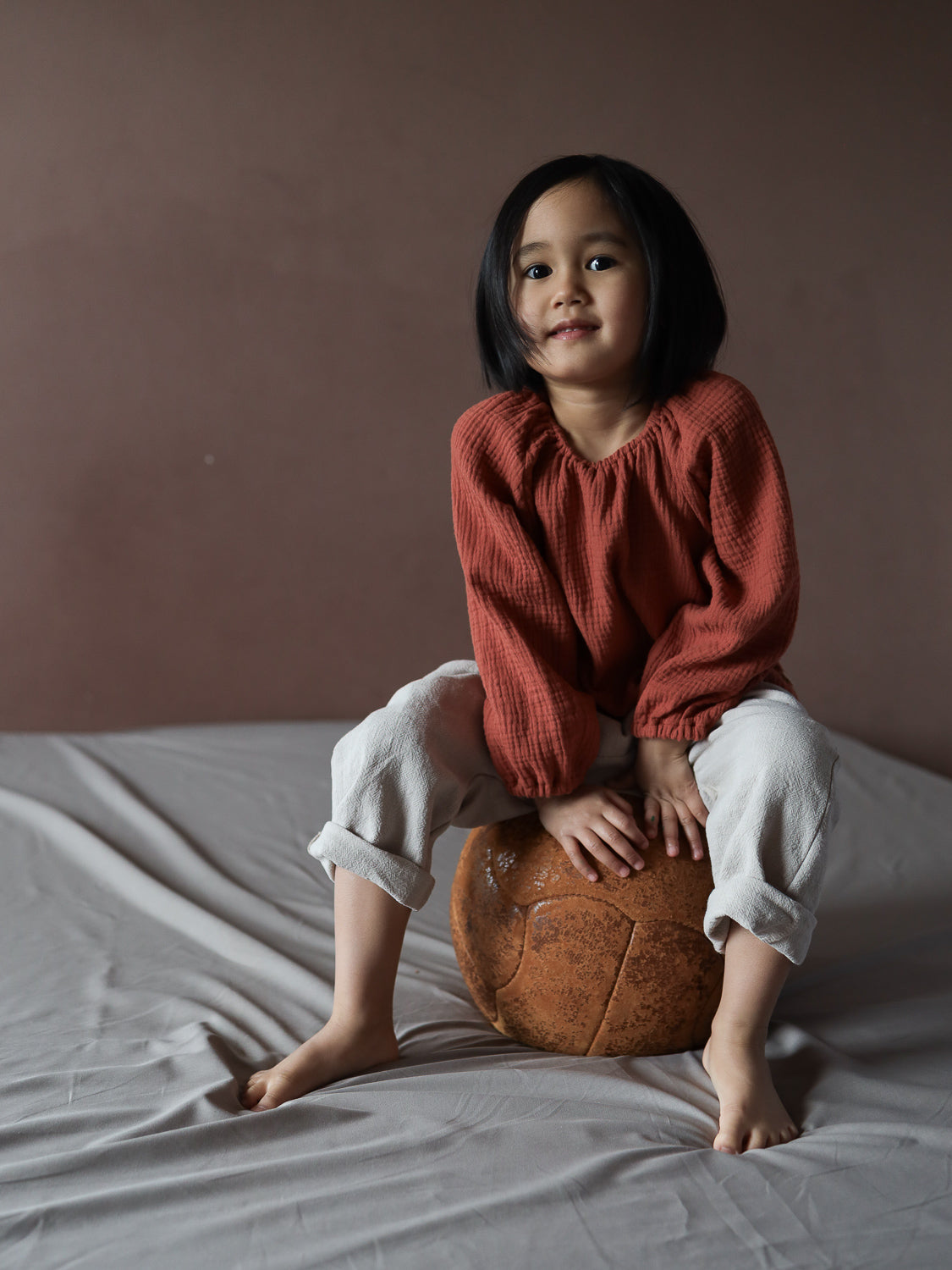 Ein Kleinkind trägt das Blüschen Musselin - Terrakotta mit lockerem Schnitt und gerafften Ärmeln, sitzt barfuß auf einem großen Lederball, zeigt den luftigen und bequemen Stil des Oberteils.
