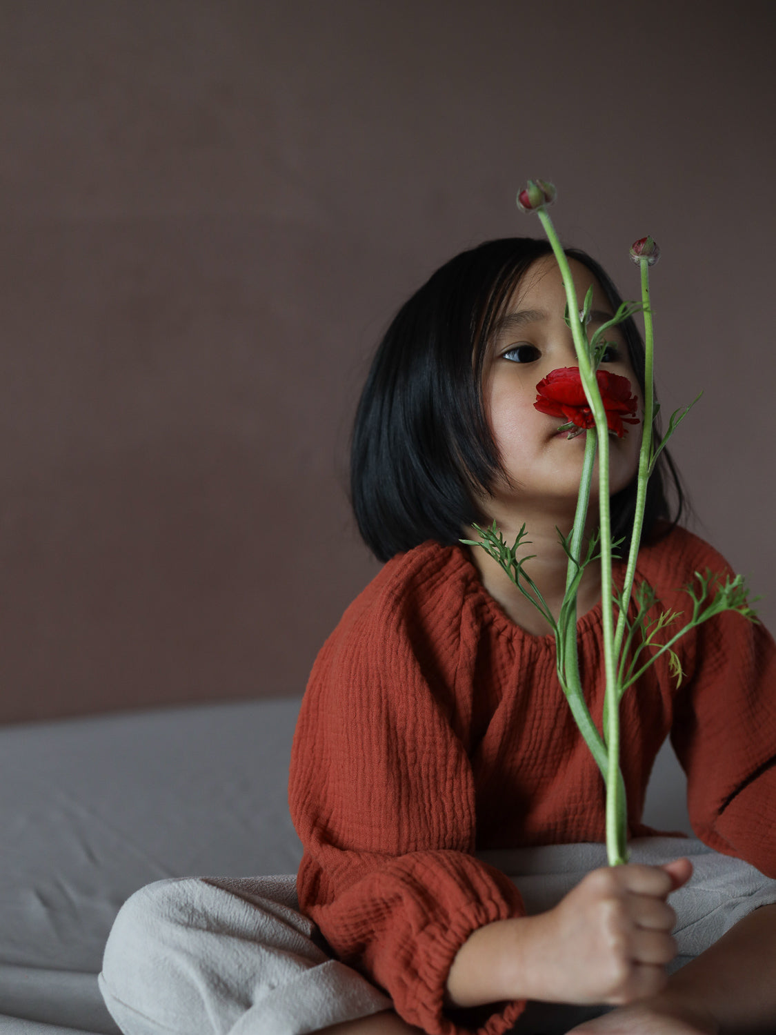 Kind hält eine Blume und trägt das Blüschen Musselin - Terrakotta, ein locker geschnittenes, strukturiertes Oberteil mit langen Ärmeln, ideal für warme Tage und empfindliche Kinderhaut.