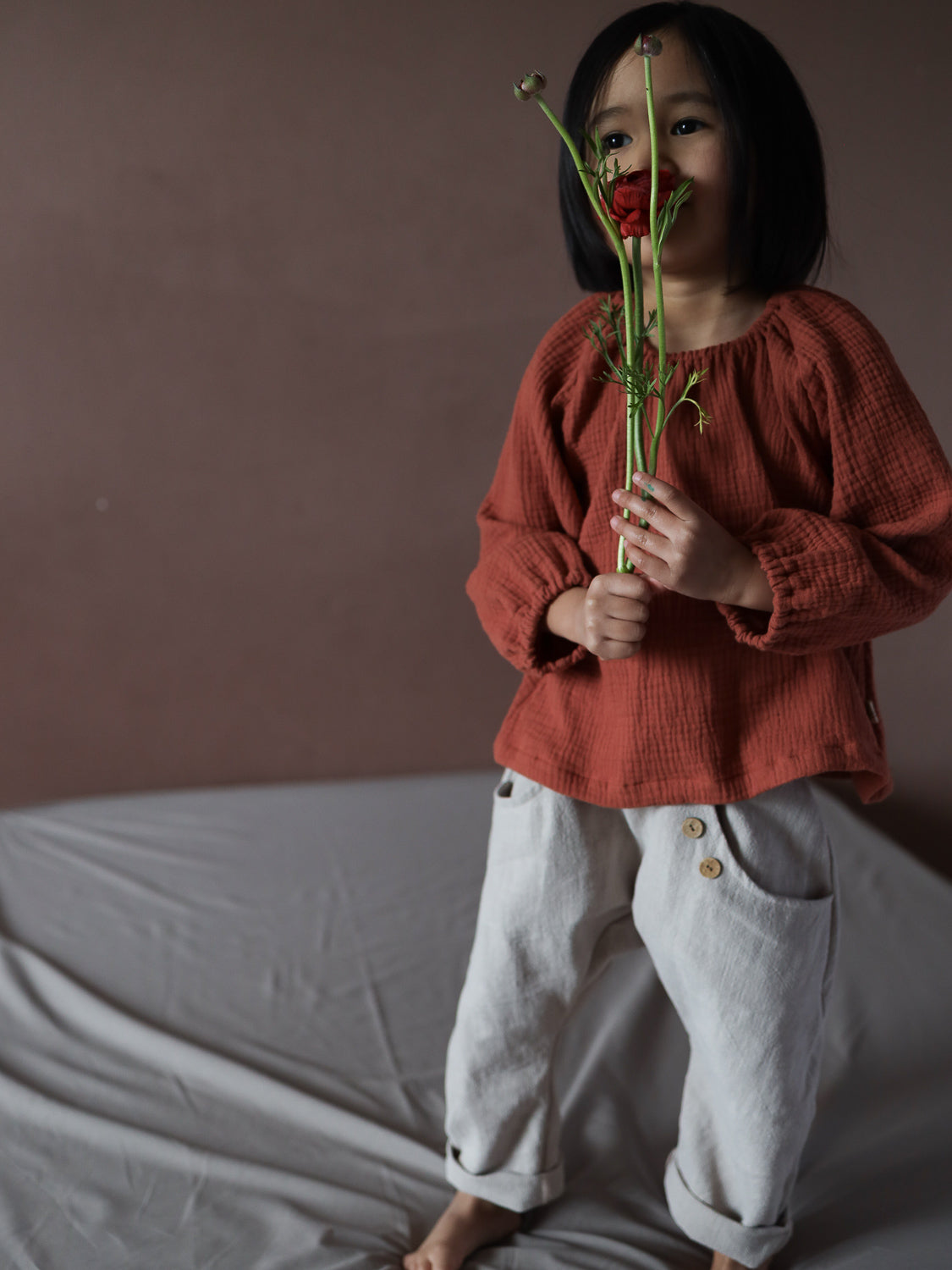 Ein Kind trägt das Blüschen Musselin - Terrakotta, ein langärmeliges, texturiertes Oberteil. Es hält eine rote Blume, trägt beige Hosen mit zwei Holzknöpfen, auf einem grauen Tuch stehend.