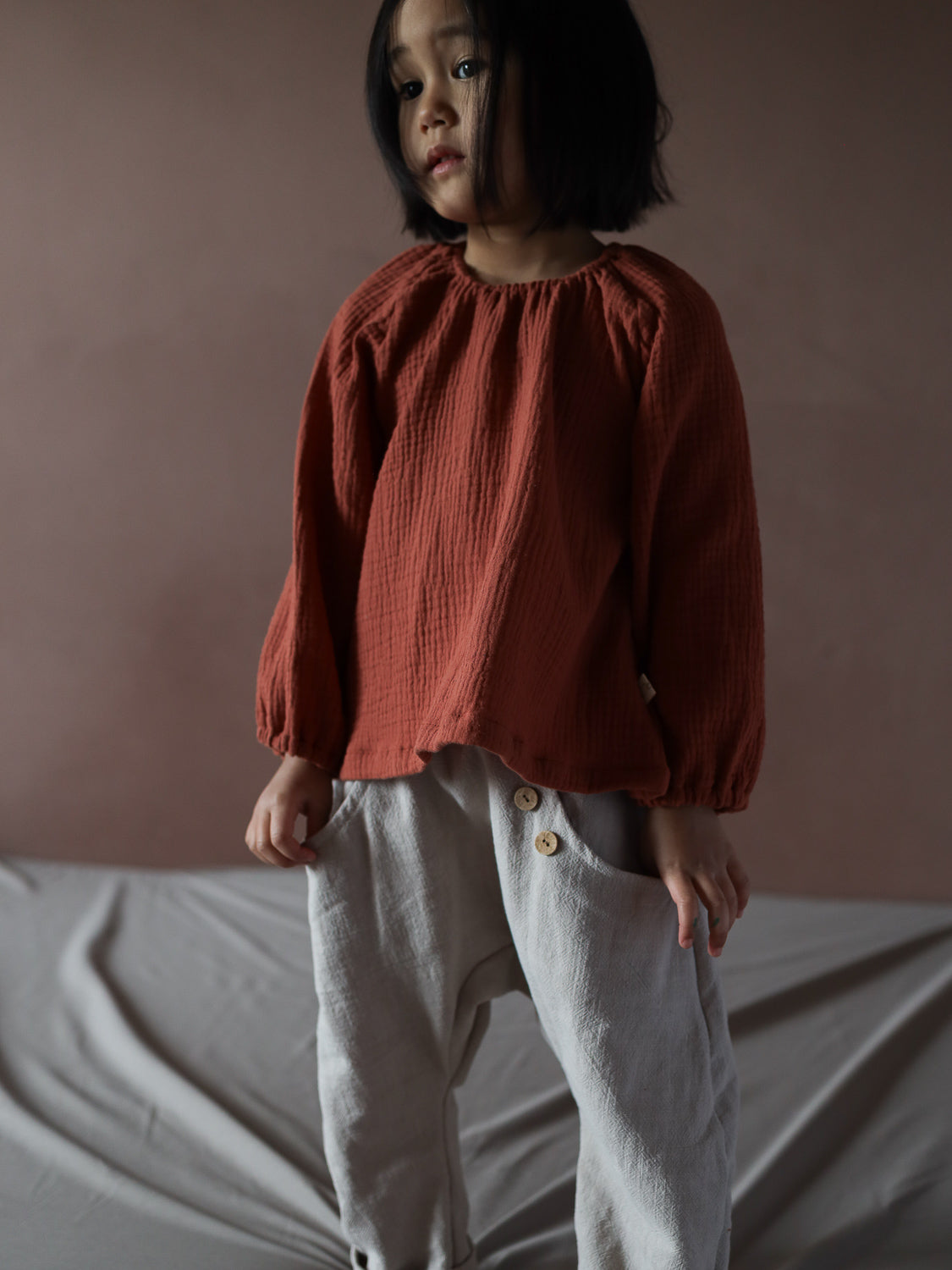 Kind trägt das Blüschen Musselin - Terrakotta, locker geschnitten mit langen Ärmeln, perfekt für warme Tage. Kombiniert mit hellgrauen Hosen, bietet es Bewegungsfreiheit und ein angenehmes Tragegefühl.