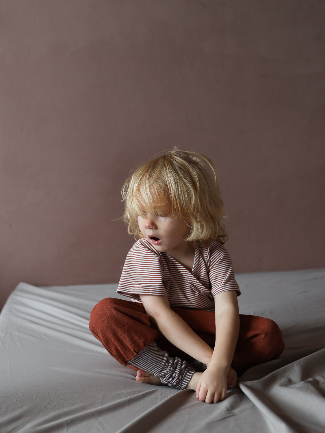 Kind mit blondem Haar trägt das Kurzes Wickelshirt Baumwolle - Ringel Ziegel, ein kurzärmeliges, gestreiftes Top, sitzt auf Bett, zeigt angenehme, weiche Passform.