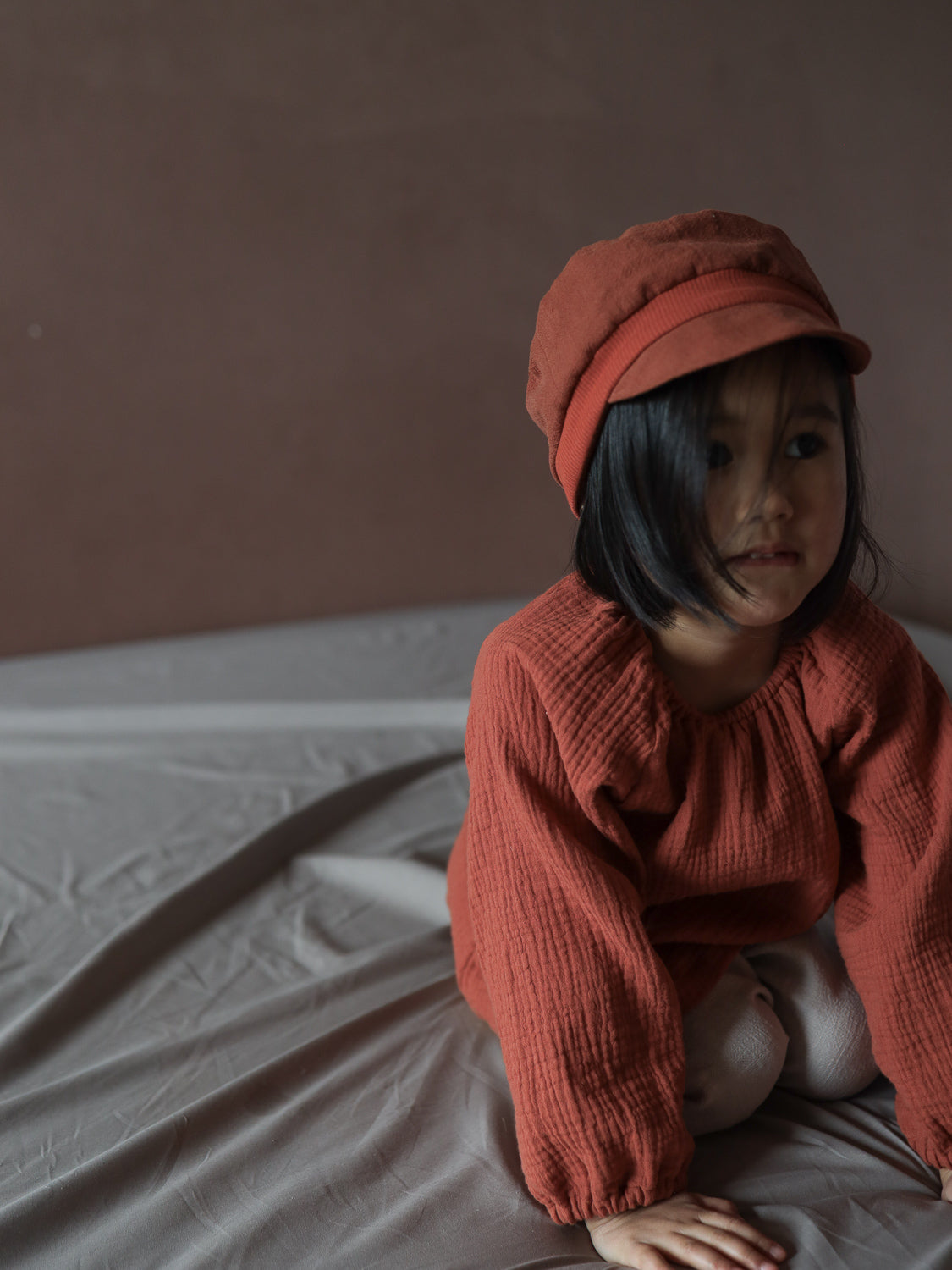 Kleinkind sitzt auf hellgrauem Tuch, trägt Blüschen Musselin - Terrakotta mit langen Ärmeln und passender Mütze. Blüschen ist locker geschnitten und ideal für warme Tage.