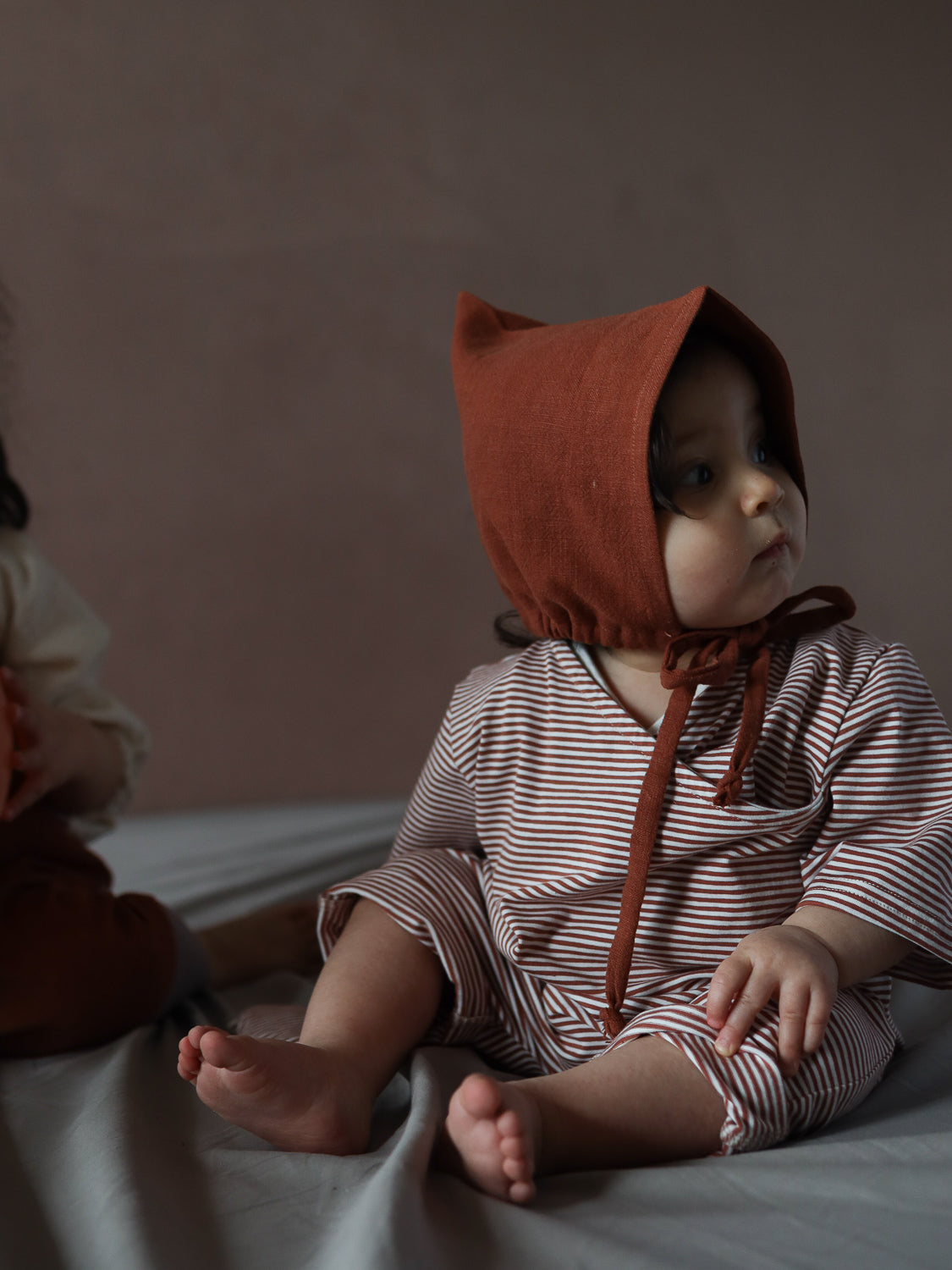 Ein Baby trägt den Wickel Overall Baumwolle - Ringel Ziegel mit rust-braunem Bonnet, sitzt auf weicher Unterlage. Der Overall ist langärmelig, gestreift und bequem, perfekt für warme Tage.