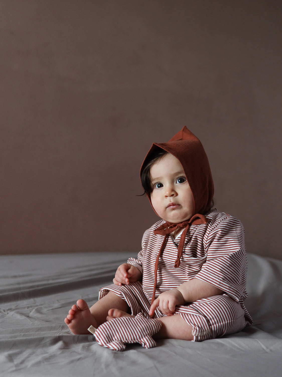 Ein Baby trägt den Wickel Overall Baumwolle - Ringel Ziegel, kombiniert mit einer passenden Haube, und hält ein gestreiftes Stoffspielzeug, das den gemütlichen und atmungsaktiven Einteiler ergänzt.