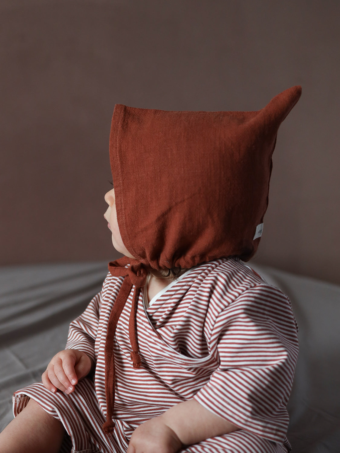 Kleinkind trägt den Wickel Overall Baumwolle - Ringel Ziegel, einen langärmligen rot-weiß gestreiften Einteiler mit praktischen Wickelausschnitt, kombiniert mit einem spitzen, rostbraunen Bonnet.