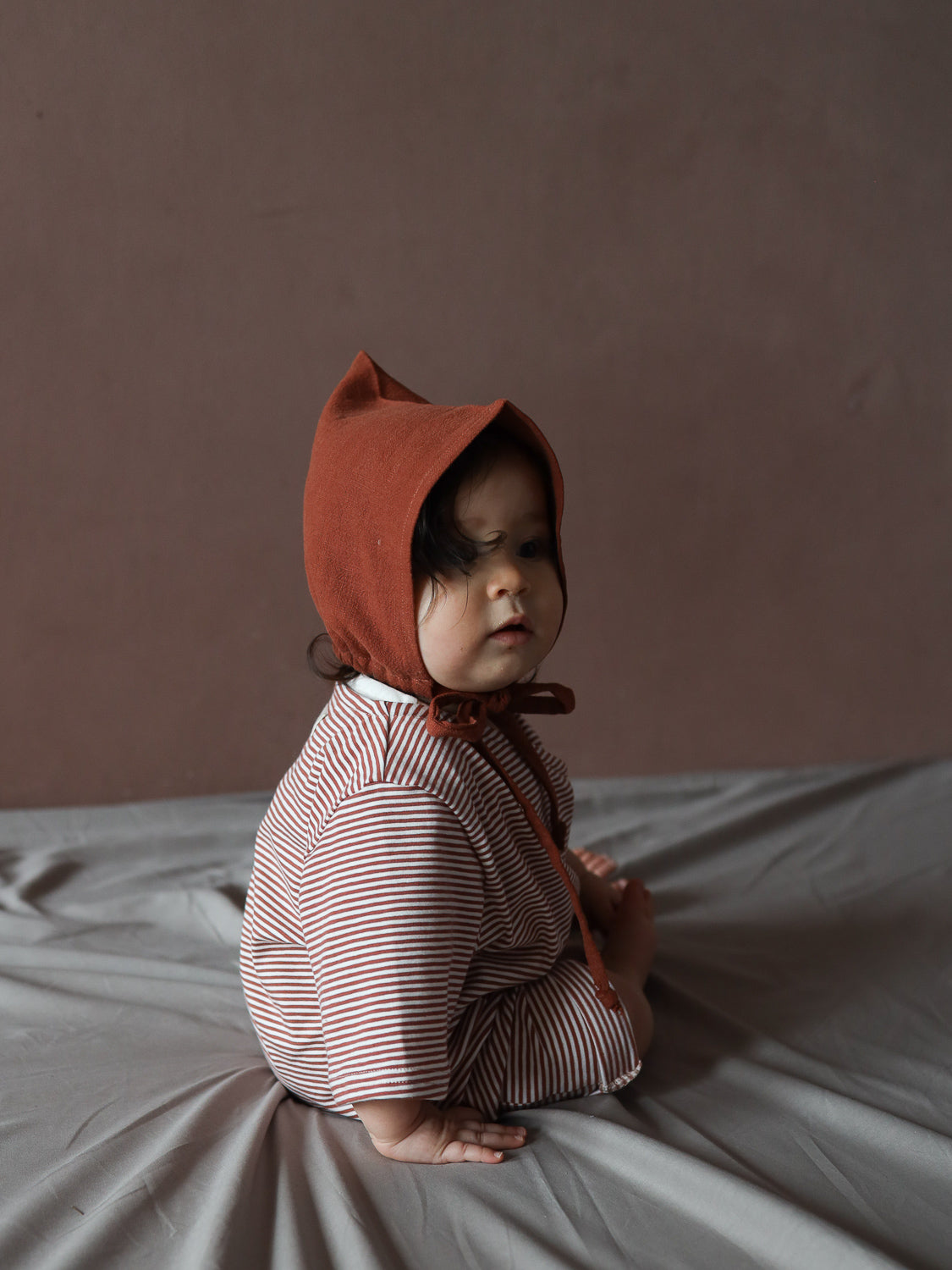 Ein Kleinkind trägt den Wickel Overall Baumwolle - Ringel Ziegel, einen gestreiften Einteiler mit praktischen Wickelausschnitt und einer Kapuze, sitzend auf einer weichen, drapierten Unterlage vor einem schlichten Hintergrund.